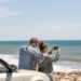 couple standing by car taking a photo of the ocean