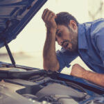 A stressed man leaning over his car’s open hood, looking at the engine.