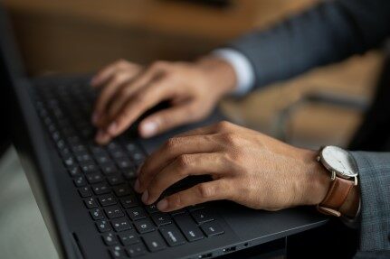The hands of a man wearing a gray plaid suit, a white shirt and a watch with a brown leather strap are shown as he types on the keyboard of a black laptop.