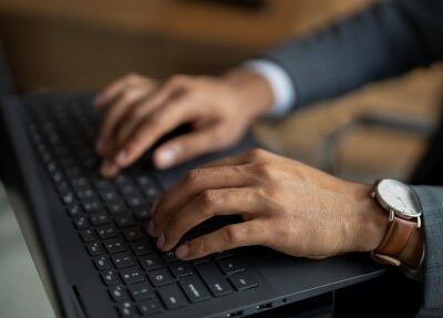The hands of a man wearing a gray plaid suit, a white shirt and a watch with a brown leather strap are shown as he types on the keyboard of a black laptop.
