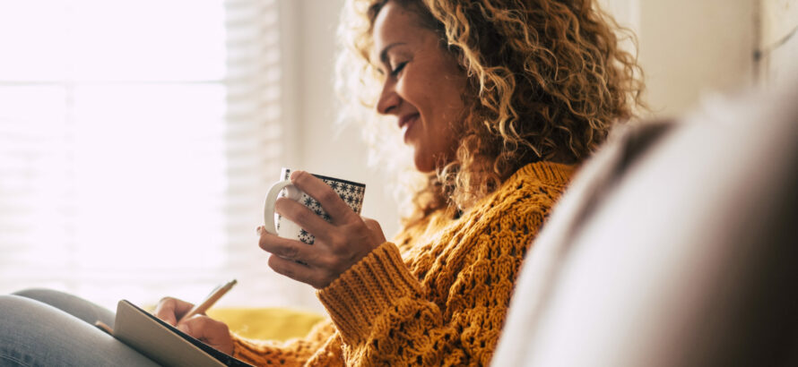 A woman writing on a journal while holding a mug.