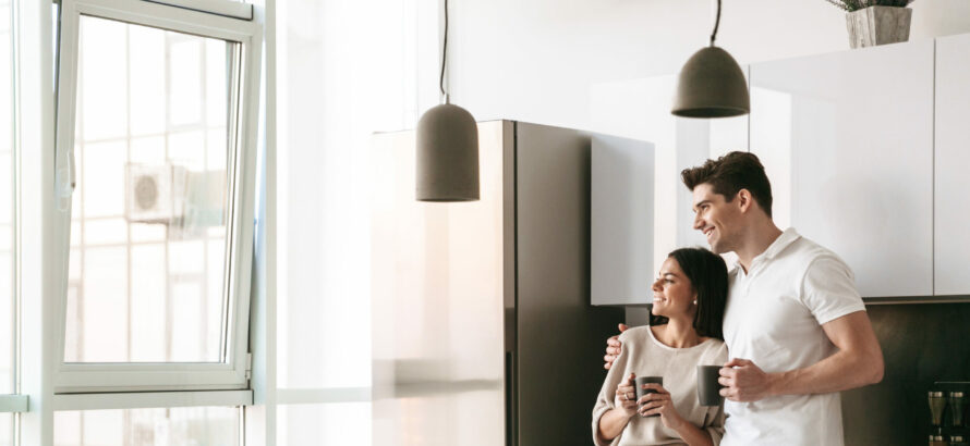 A couple looking out the kitchen window while holding mugs in their hands.