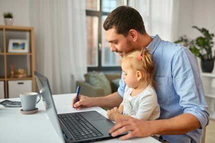 A man writes on paper while holding a baby in front of a laptop.