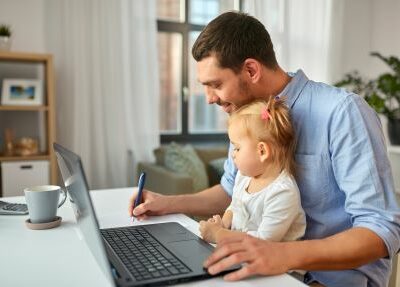A man writes on paper while holding a baby in front of a laptop.