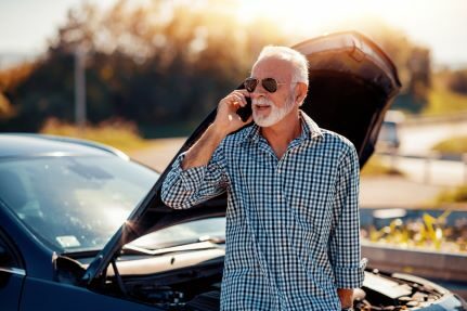 A man making a phone call in front of a car with the hood open.