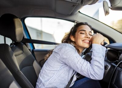A woman smiles while leaning on her steering wheel.