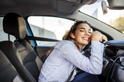 A woman smiles while leaning on her steering wheel.