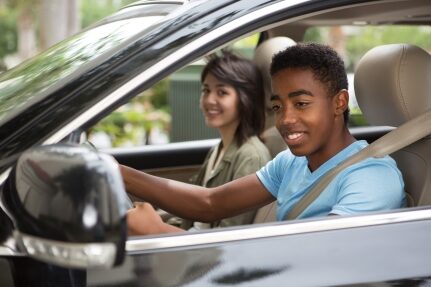 A teen drives a car while a woman sits smiling in the passenger seat.