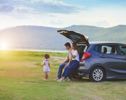 A couple sits in the back of a car parked in an open field watching their child.