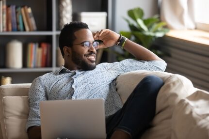 A man smiles while sitting on a couch with a computer in his lap.