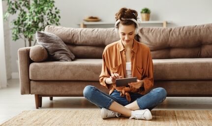 A woman looks at her tablet while sitting in front of a couch on the floor.