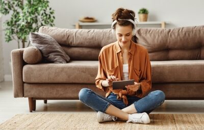 A woman looks at her tablet while sitting in front of a couch on the floor.