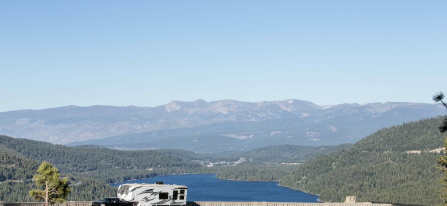 An RV driving on a scenic road; a tree-lined lake and mountains appear in the background.