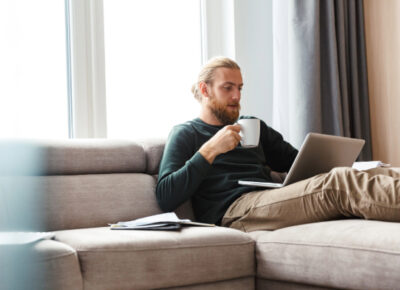 A man sits on his couch, holding a cup of coffee and looking at his laptop.