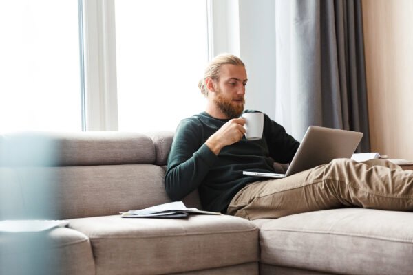 A man sits on his couch, holding a cup of coffee and looking at his laptop.