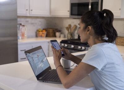 A woman using her smartphone and laptop in the kitchen.