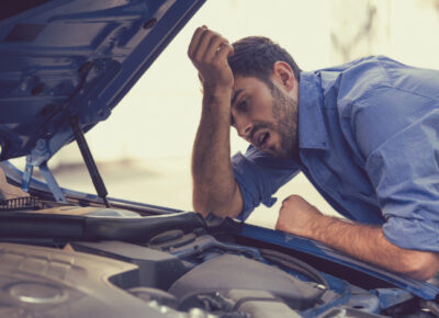 A stressed man leaning over his car’s open hood, looking at the engine.