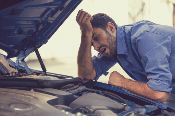 A stressed man leaning over his car’s open hood, looking at the engine.