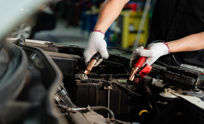 A mechanic, wearing gloves, puts black and red jumper cables to a car battery.