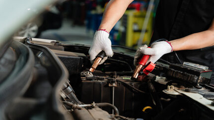 A mechanic, wearing gloves, puts black and red jumper cables to a car battery.
