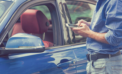 A man, standing outside of his blue car, checks his smartphone.