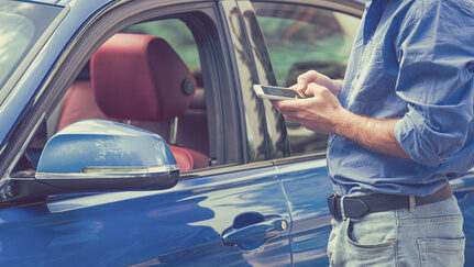 A man, standing outside of his blue car, checks his smartphone.