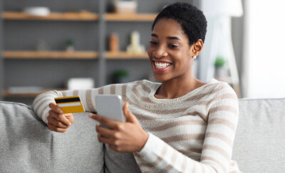 A woman happily holds a credit card in one hand and her mobile device in her other hand.