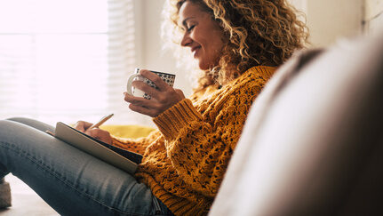 A woman sits on her couch while holding a mug and writing in her notebook.