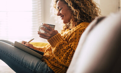 A woman sits on her couch while holding a mug and writing in her notebook.